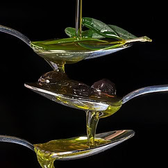 Se vierte aceite sobre tres cucharas apiladas, con la cuchara superior sosteniendo una hoja y la cuchara del medio con aceitunas negras. El aceite de oliva andaluz cae en cascada hasta el fondo de la cuchara sobre un fondo negro.